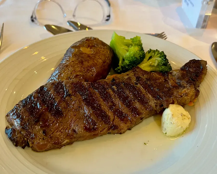 A white plate with a charred steak, baked potato, broccoli and garlic butter