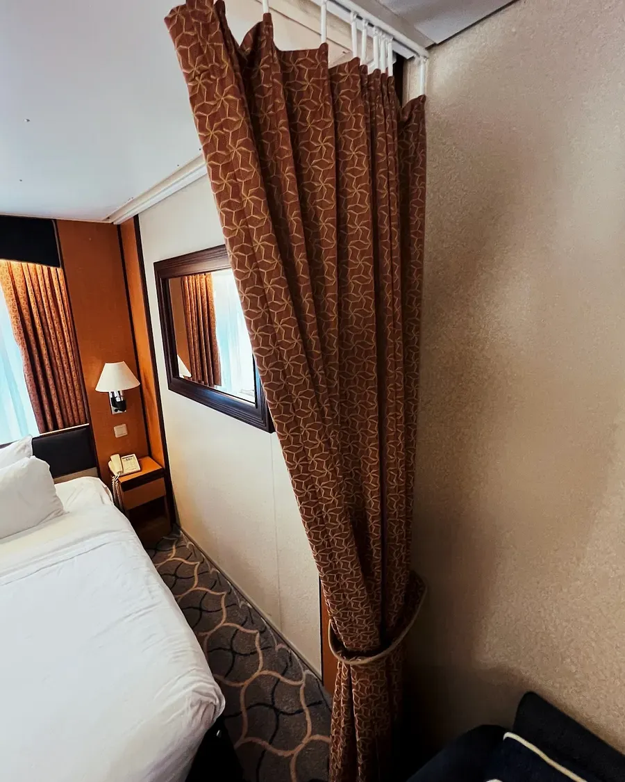 A heavy curtain is shown which separates an ocean view cruise liner cabin bed from the rest of the room