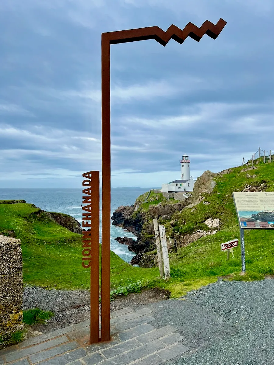 Rusted metal Fanad Head lighthouse marker with white lighthouse on rocky coastal cliffs in background