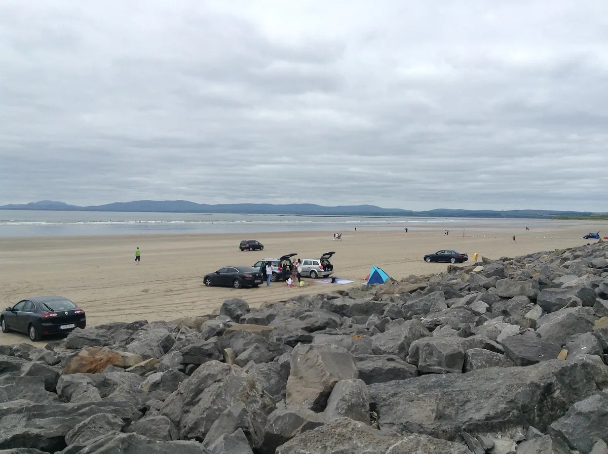 Wide sandy beach at Rossnowlagh with cars parked on sand, rocky breakwater in foreground, distant mountains