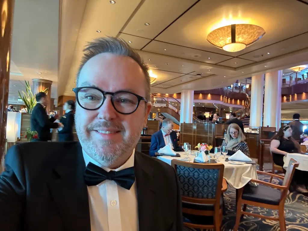 Man in formal attire with bow tie and glasses taking selfie in cruise ship dining room
