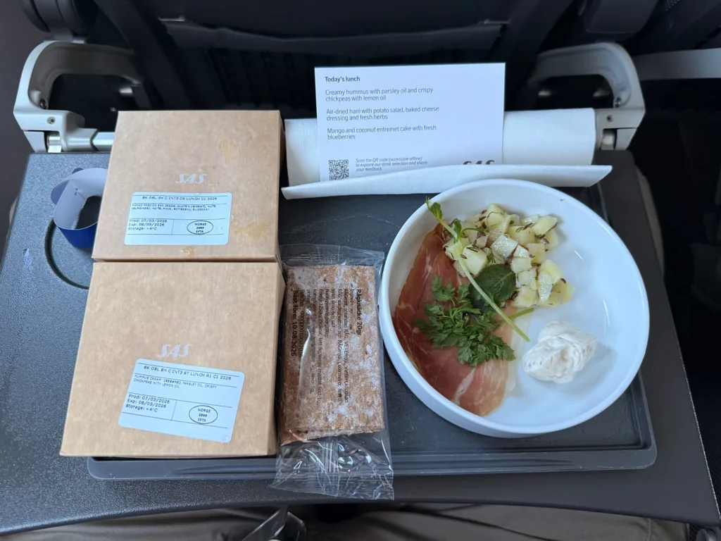 Airline meal tray with prosciutto, potato salad, bread, and two boxed items on a fold-down table