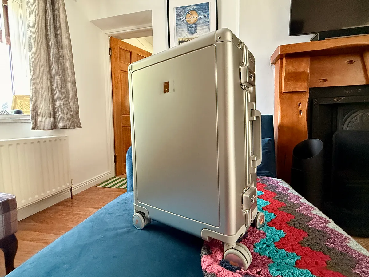 Silver hardshell suitcase on wheels in a living room with fireplace and colorful rug