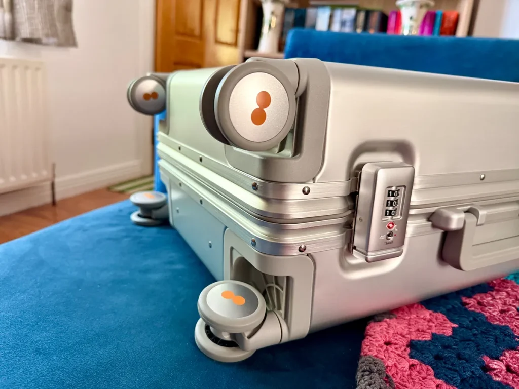 Silver hardshell suitcase with orange accent wheels and combination lock on blue bed