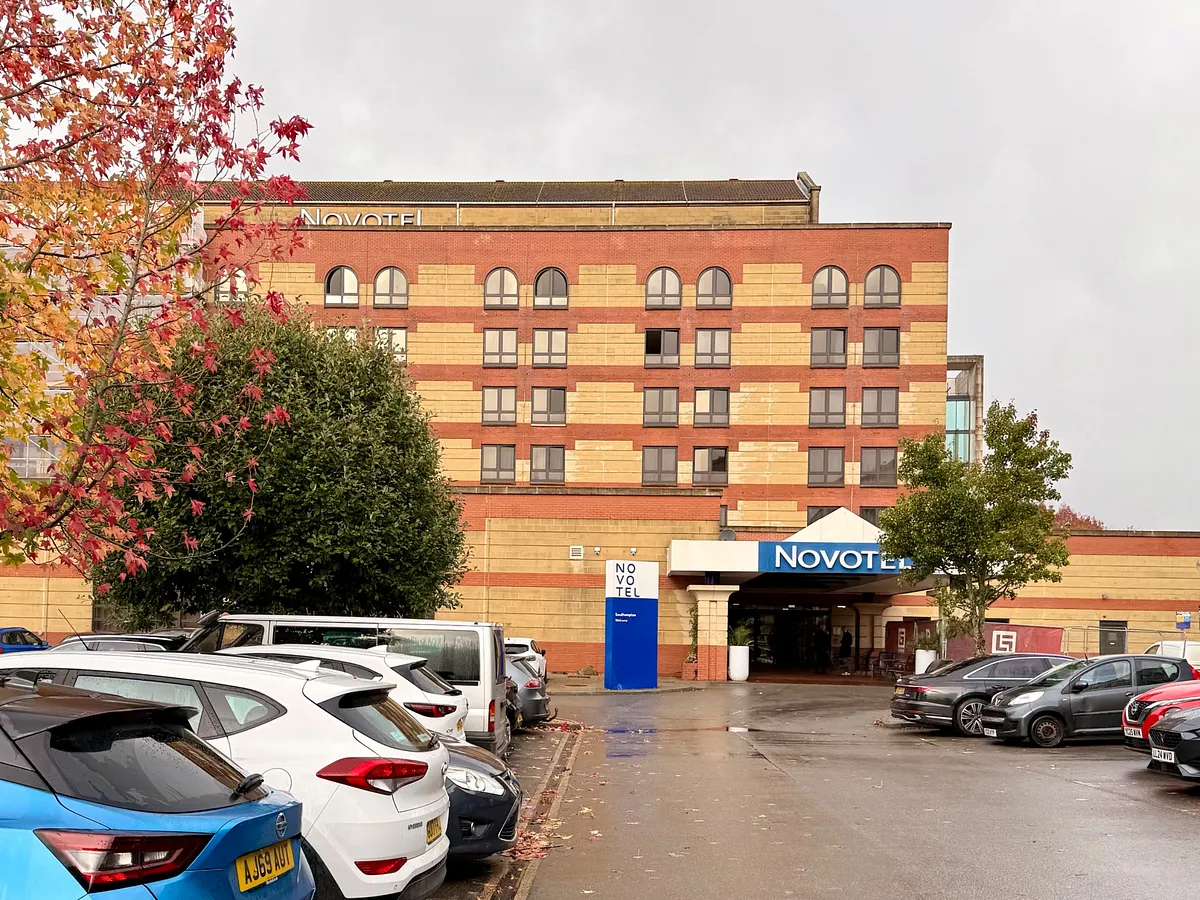 Novotel hotel exterior with red brick and yellow facade, parking lot with autumn trees in foreground